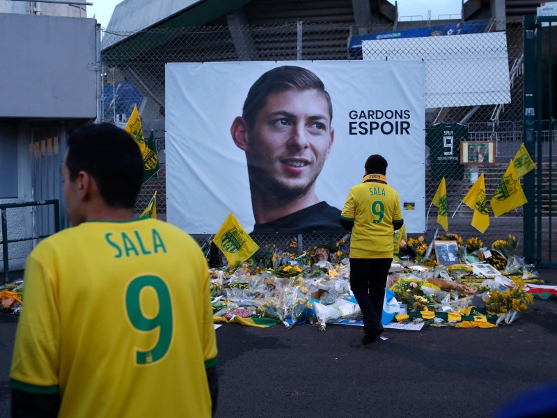 A man in a yellow football shirt with the number 9 and the word Sala on it stands in front of a tribute to a dead football player.