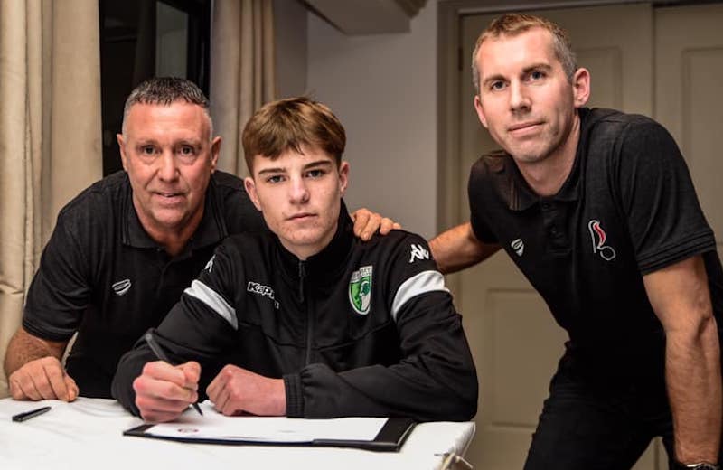 A young man looks a the camera, as he signs his first professional football contract. Two older men either side smile and look at the camera.