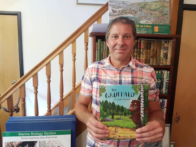 A man - Yan Marquis - holding a copy of the Gruffalo translated into Guernsey French. He stands in front of a bookshelf and some stairs.