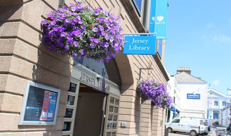 A stone building with arched entrances displays a blue sign reading “Jersey Library” above the doorway. Two large hanging baskets overflow with bright purple flowers on either side of the entrance. A poster frame is mounted beside the door. In the background, a street with shops, signs, vehicles, and a blue sky is visible.