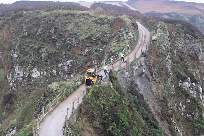 La Coupée has been closed to all traffic this week while the damage was repaired. One worker hangs using ropes and a harness over the sheer drop, inspecting the concrete. A small JCB Forklift takes up the majority of the path. 