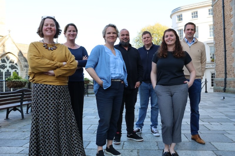 A group of seven people - four women and three men - stand in a paved courtyard surrounded by historic stone buildings. They are dressed in casual and smart‑casual clothing, with benches and greenery visible in the background on a sunny day.