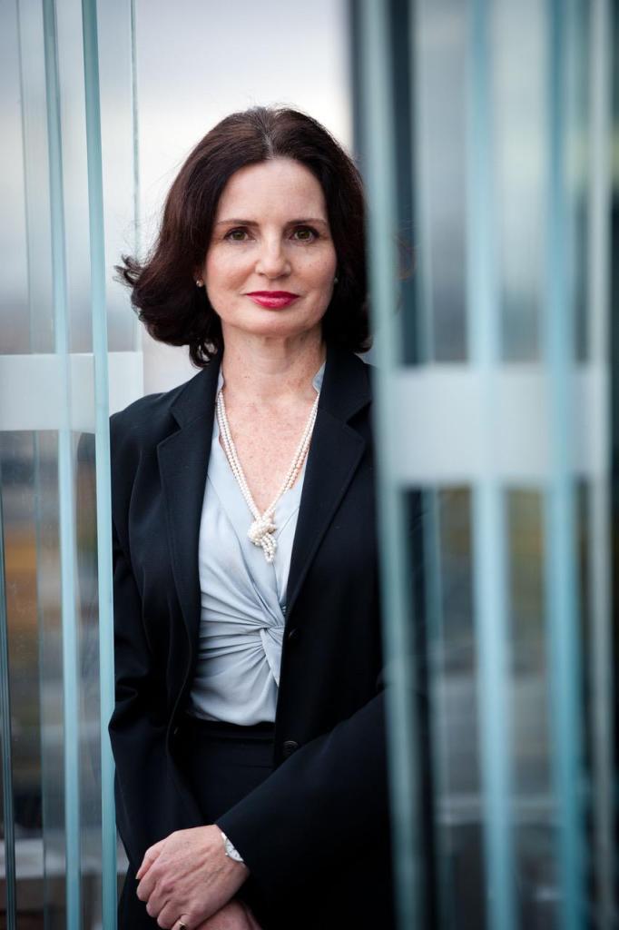 A woman with shoulder length brown hair in a business suit looks at the camera, surrounded by corporate glass.