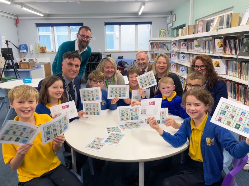 A group of primary school children sit around a table in a library holding up laminated Challenge Cards. Several teachers and deputies stand behind them.