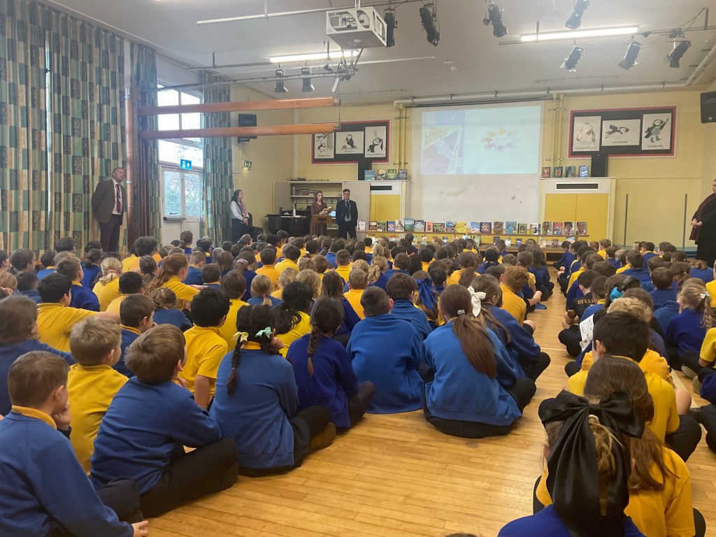 More than 100 primary school children in blue and yellow uniforms sit on a wooden school hall for an assembly on reading.