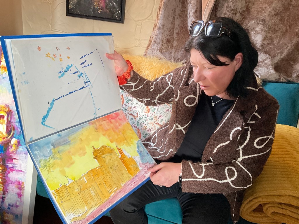 A woman with shoulder-length brown hair in a brown jacket shows a watercolour sketch of Elizabeth College - an old private school building.