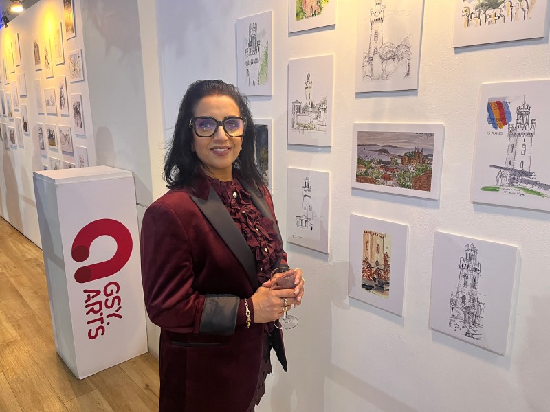 A woman with glasses in a purple jacket stands in front of a selection of sketches in a gallery.