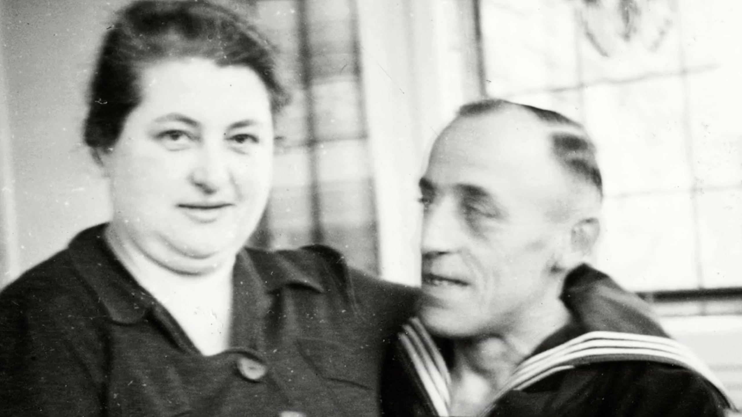 A black and white photo of a large woman in a dark blouse looking at the camera. Her husband, dressed in a sailor's uniform, looks at her lovingly.