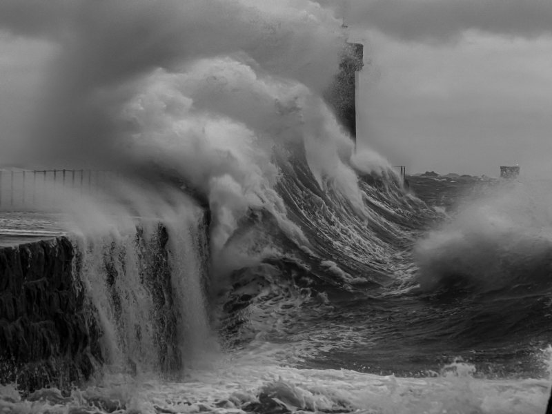 A black and white photo of a large wave breaking over the St Peter Port lighthouse.