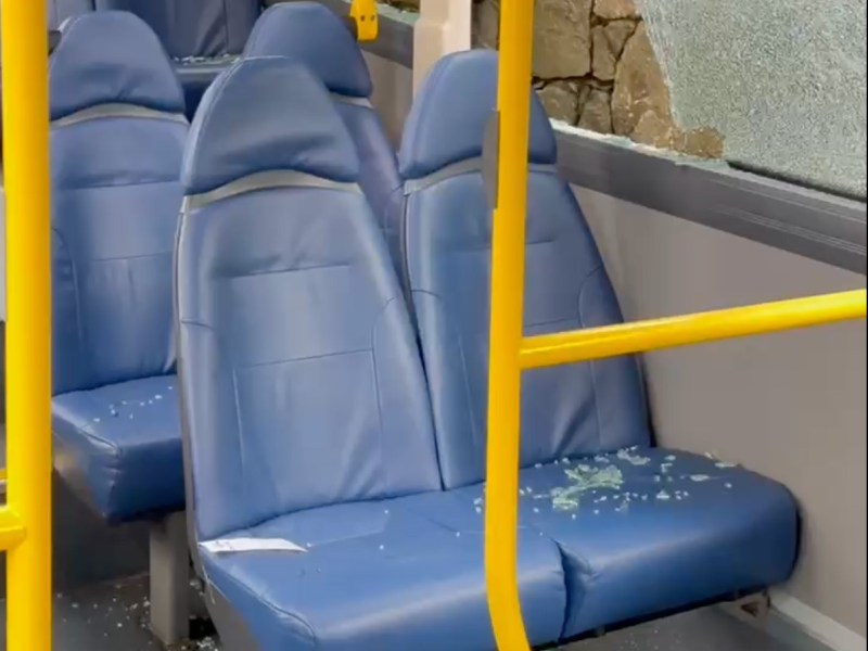 A smashed bus window, with glass all over the seat and a granite wall outside.