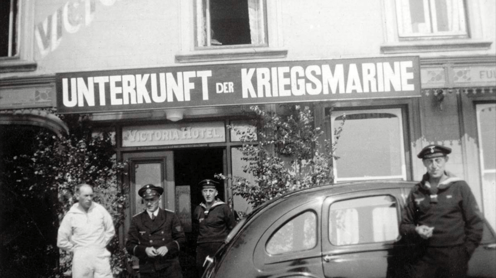 A black and white photo of the Victoria Hotel with a sign in German above which reads Unterkunft de Kriefsmarine. Several troops and officer stand in front. One leans on a VW Beetle smoking.