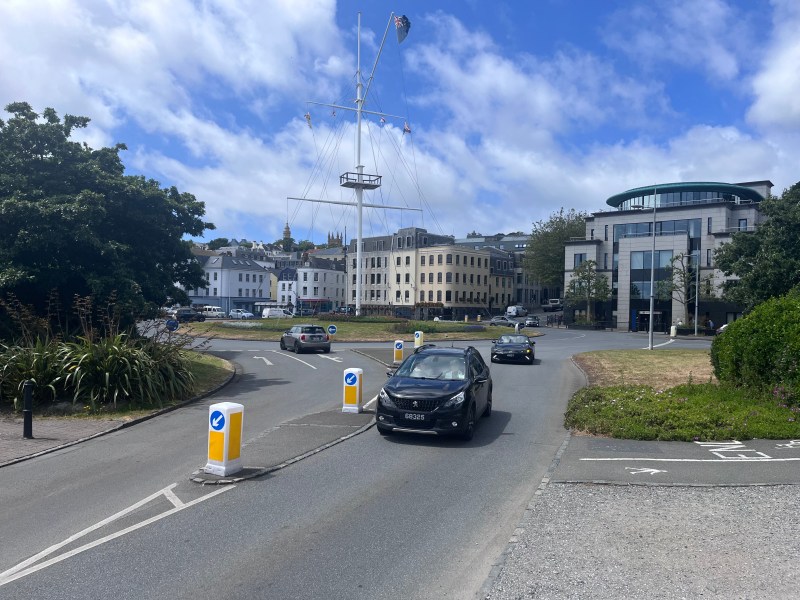 A car pulls off a roundabout on a sunny day. The roundabout has a large flagpole in the middle, and modern corporate buildings on one side, with a bush on the other.