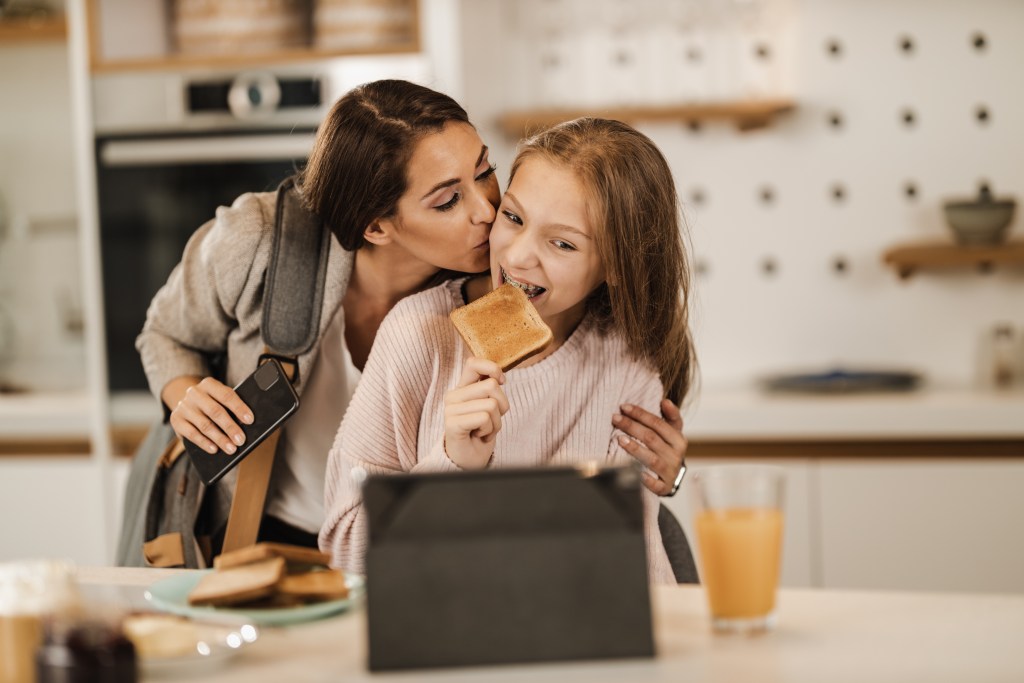 A young mother kisses her teenage daughter, who is eating toast and playing with an iPad.