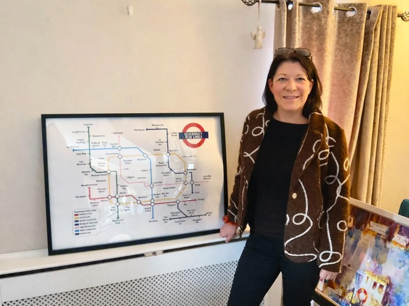 A woman in a brown jacket smiles as she stands in front of a London-underground-style map of Guernsey pubs.