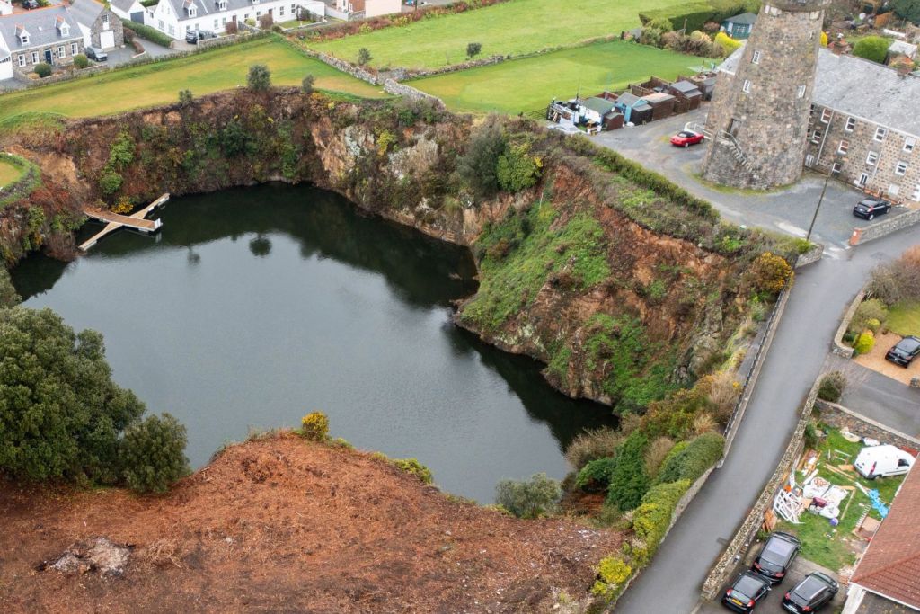 Aerial view of a deep, water-filled quarry surrounded by steep rocky cliffs with patches of greenery, located beside a stone tower and nearby houses, roads, and parked cars.
