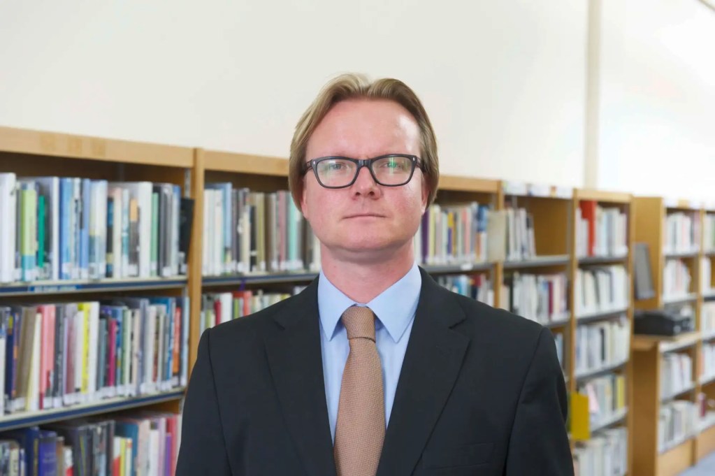 A bespectacled man in a suit stands in a library.