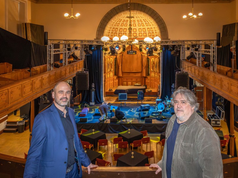 Two men in suit jackets stand facing each other with a wood-panelled arts venue behind them.