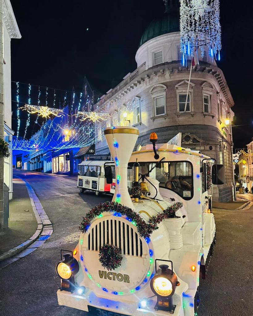 A small white tourist road train decorated with colourful Christmas lights and tinsel is parked on a street at night. Festive light displays hang overhead, illuminating the curved stone building behind it and the quiet, gently sloping street lined with shops.
