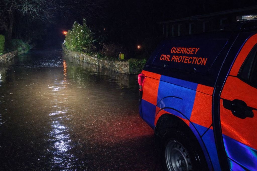 A Guernsey Civil Protection vehicle illuminates a flooded rural road at night, with water covering the roadway and stone walls visible along the sides in the darkness.