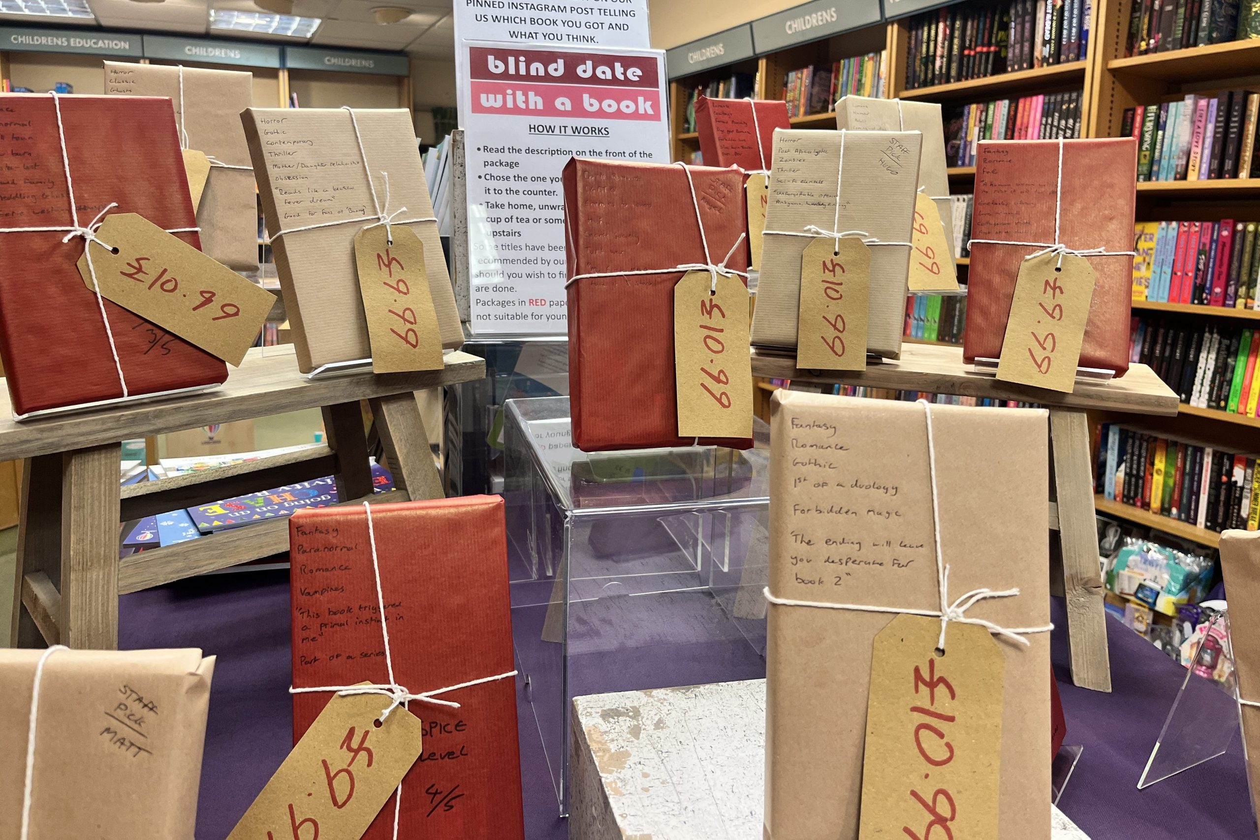A bookshop display shows wrapped books in brown and red paper tied with string, arranged on small stands as part of a blind date with a book promotion. Each book has a large handwritten tag, and shelves of colourful books fill the background.