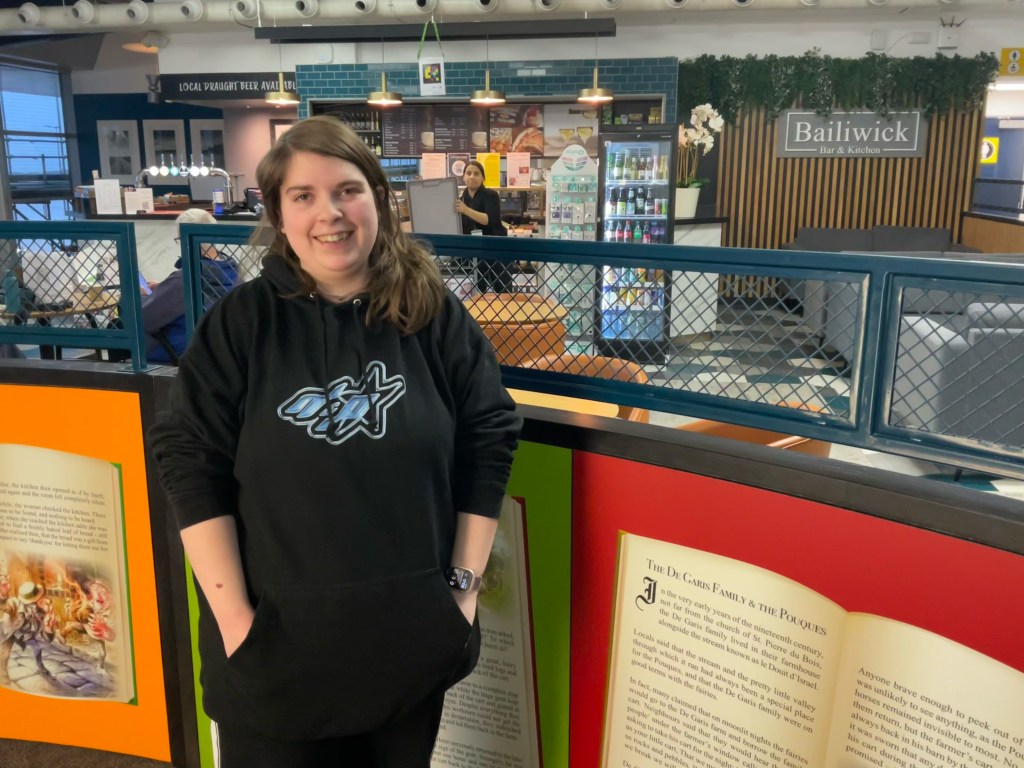 A woman wearing a black hoodie with a stylized graphic on the front stands indoors with her hands in her pockets. She is positioned in front of a colourful display featuring large panels designed to look like open book pages. Behind them is a café-style area with hanging lights, a counter, refrigerated drink cases, and a sign that reads Bailiwick Bar & Kitchen. Another person is visible working behind the counter. The setting has a modern, casual atmosphere with teal accents and greenery.
