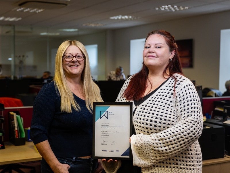 Two women stand together in an office workspace, holding a framed certificate between them. One wears a dark long‑sleeved top and has long light‑colored hair, while the other wears a light, open‑knit sweater over a dark top and has long reddish hair. Behind them, office desks, chairs, computers, and bright windows create a busy indoor setting with overhead lighting.