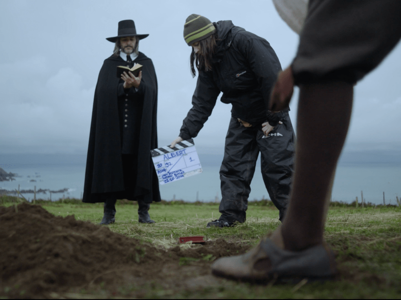 An actor dressed in a historical black puritan's outfit and buckled hat, holding up a small black book - he stands in a field on a cliff. A film crew member holds a clapper board in front which says: Albert. The shoe and leg of another actor are in shot at the front.