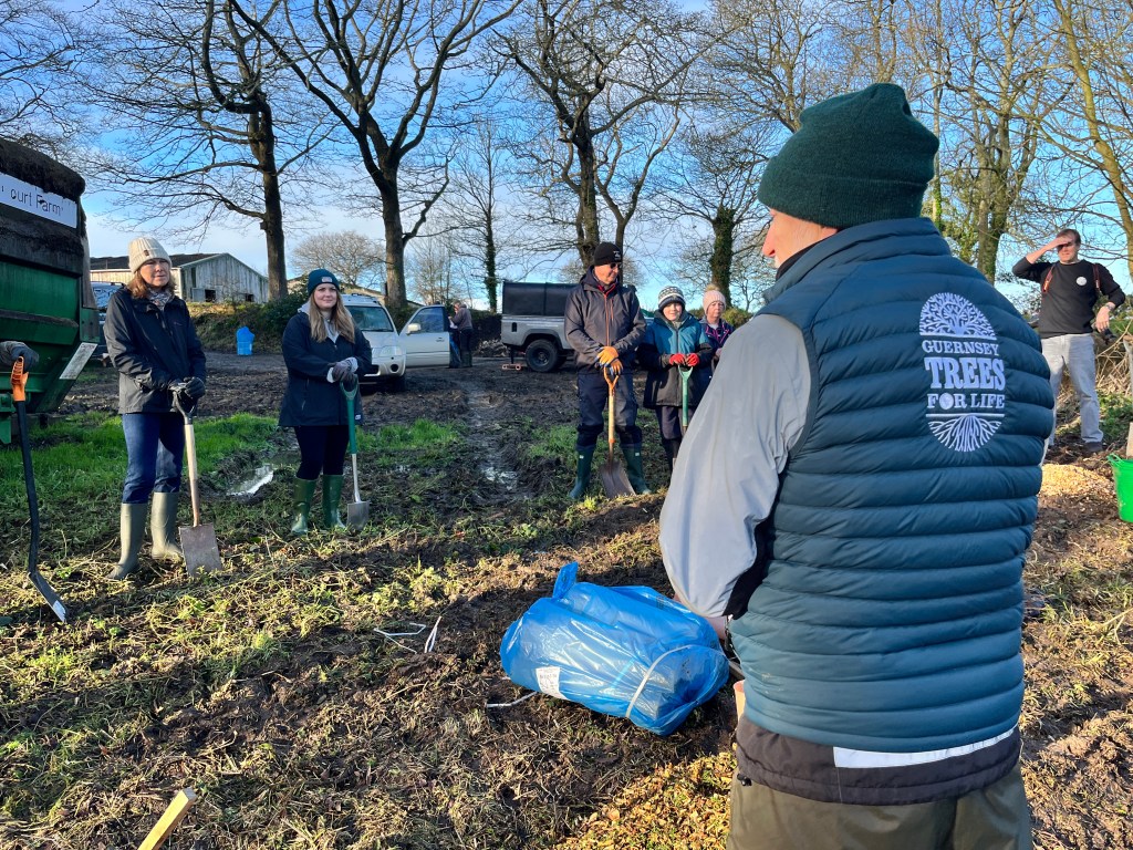 A man in a teal Guernsey Trees for Life bodywarmer and a green beanie hat talks to spade-wielding volunteers in a field in Guernsey.