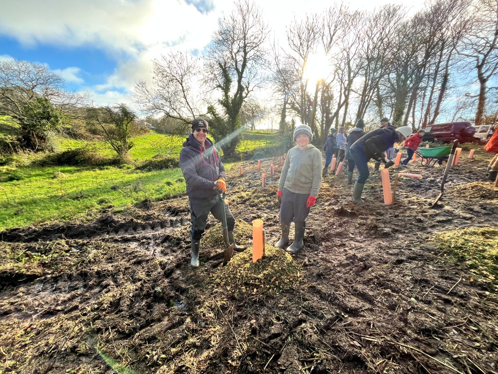 A man and a young boy smile as they stand in a muddy field next to a tree they've just planted.