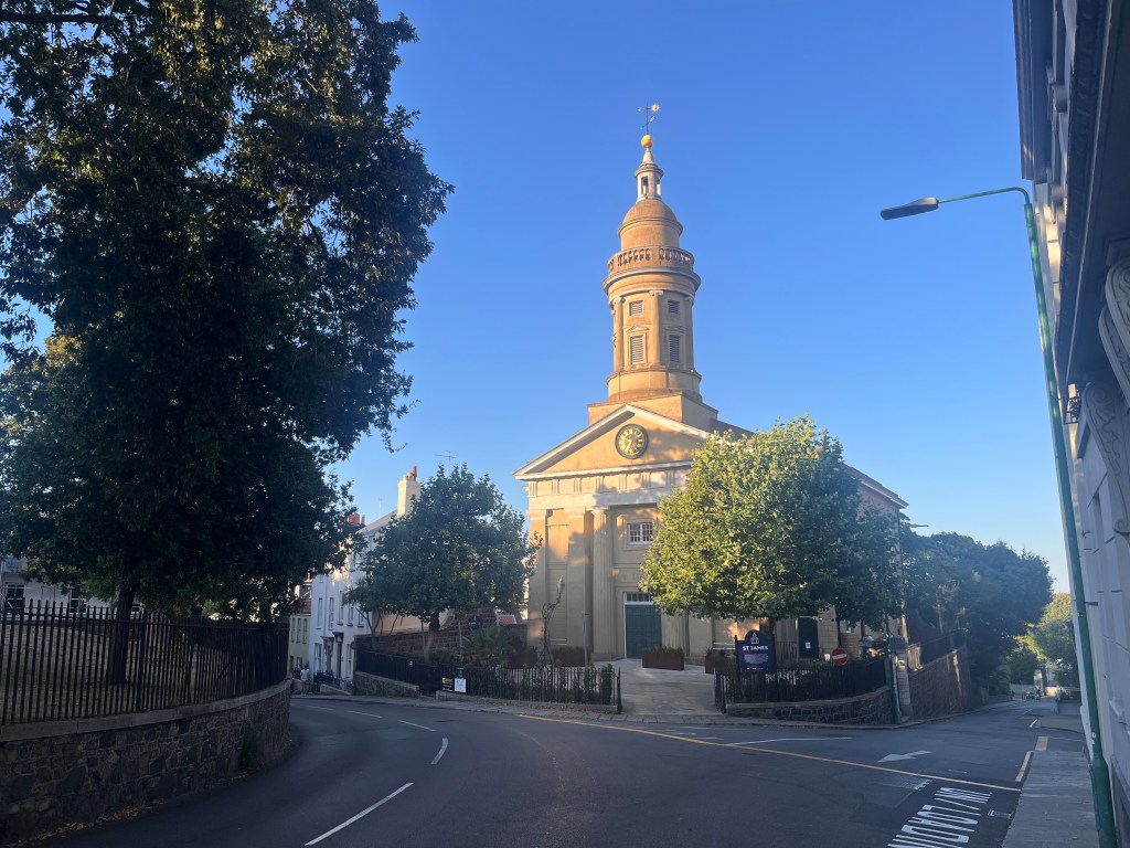 A historic church building - now an arts centre - with a tall clock tower and spire stands at a quiet street corner under a clear blue sky. The sun lights up the pale stone facade, while trees frame the building on both sides and a curved road runs in front.
