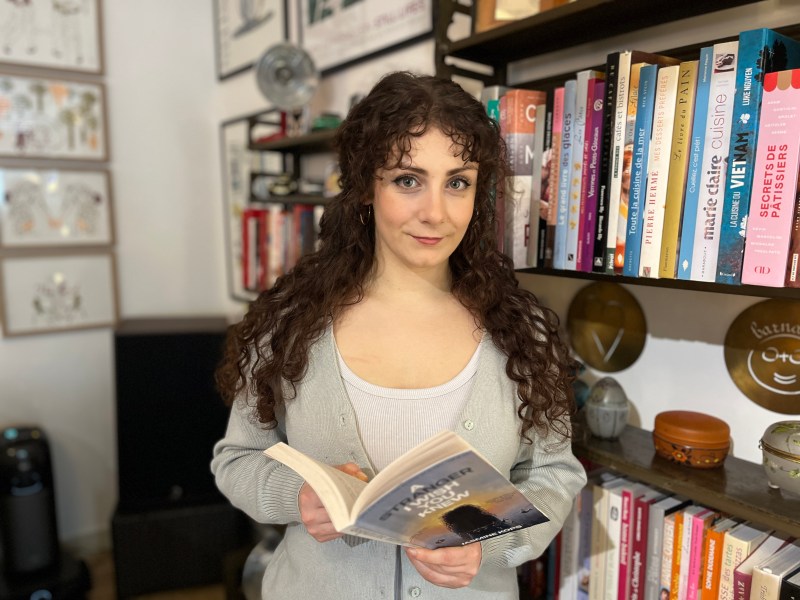 A smiling woman with long, dark, curly hair stands beside a bookshelf filled with colourful books - she is holding an open copy of a book called 'A Stranger I Wish I Knew' by Jasmine Kops. She is wearing a light cardigan over a white top. The setting appears to be a home library, with additional shelves and objects visible in the background.