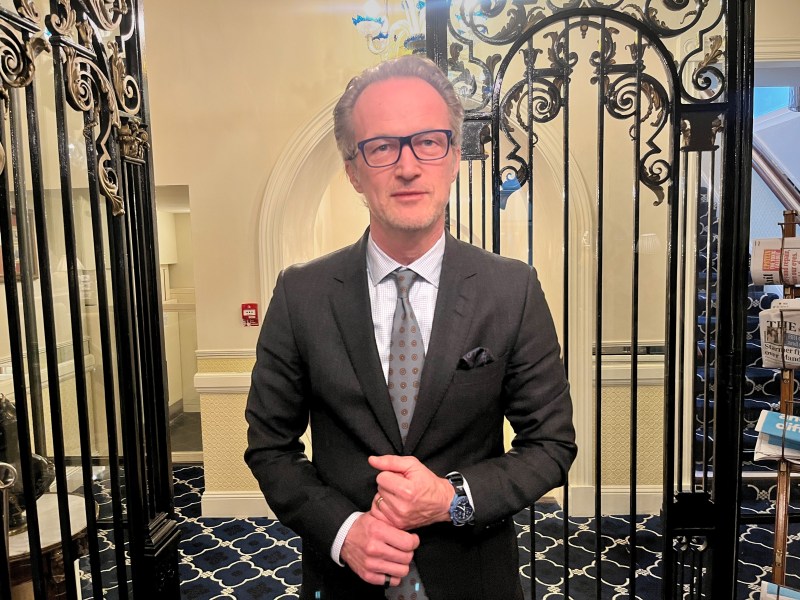 Christian Gattiker . A man with grey hair and glasses in a dark blue suit, white shirt and blue tie. He stands in front a decorative glass door in a hotel.