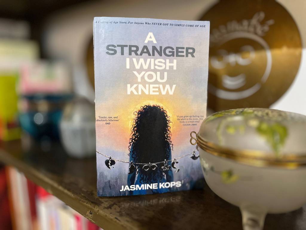 A book titled 'A Stranger I Wish You Knew' by Jasmine Kops is displayed standing upright on a wooden shelf. The cover shows a silhouetted figure with curly hair looking toward a sunrise or sunset sky, with string lights hanging across the bottom of the scene. The background includes blurred household objects and décor on the shelf, including bowls and containers.