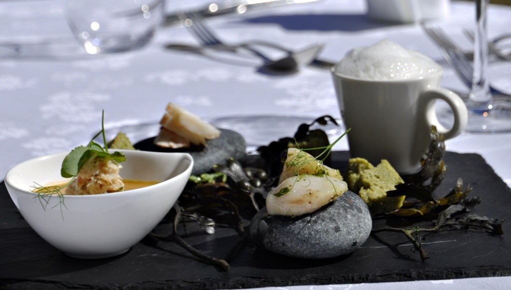 A close-up of a seafood tasting plate on a black slate, featuring prawns, seaweed, a small bowl of soup, green savoury bites and a foamy drink in a white cup on a white tablecloth.