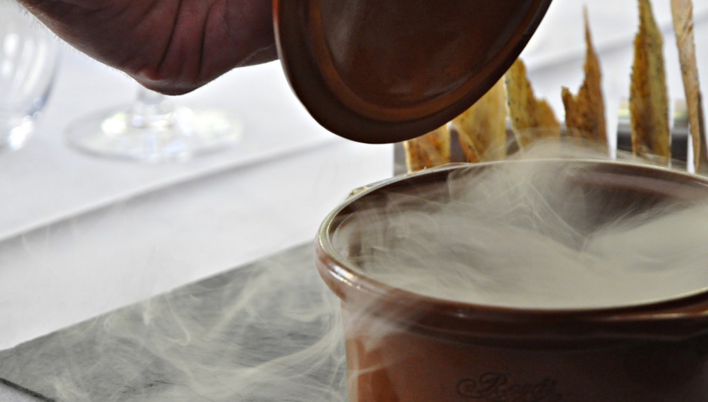 A hand lifts the lid from a small brown pot as white steam billows out, with crisp bread pieces standing upright behind it on a white tablecloth.