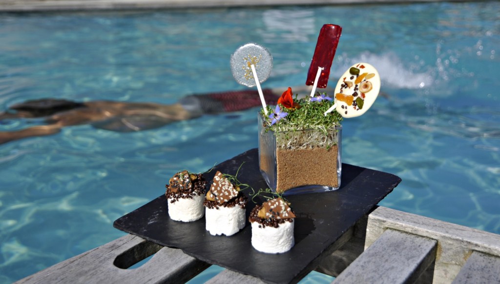 A slate board holding three small decorated desserts sits beside a swimming pool, with a glass jar filled with soil‑like crumbs, edible flowers and lollipops behind them. A swimmer moves through the water in the background.