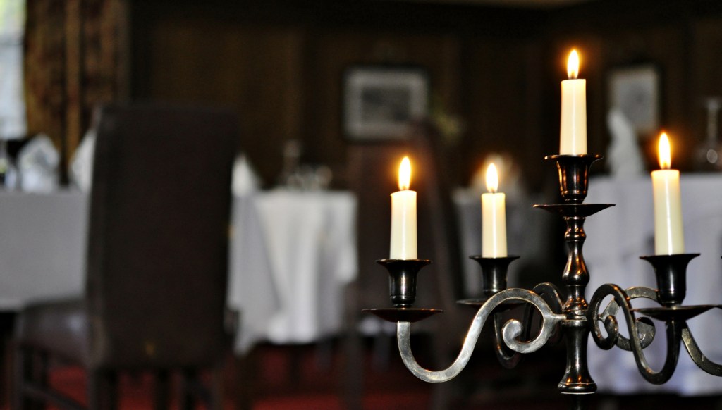 A metal candelabra with lit candles stands in the foreground of a softly lit dining room with white tablecloths and dark wood-paneled walls.