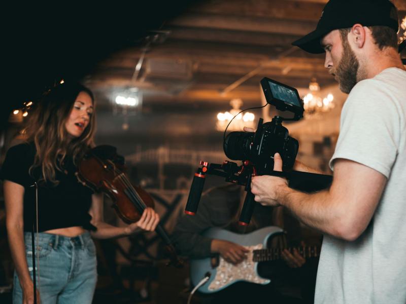 A person operating a professional video camera films a musician playing a violin in an indoor setting with warm, low lighting. Another musician holding a guitar is partially visible in the background. The scene appears to be part of a music video or live performance recording, with camera equipment and soft overhead lights contributing to a studio-like atmosphere.