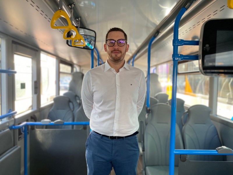 A man standing inside a modern bus, facing the camera with hands behind their back. He is wearing a white long‑sleeve button‑up shirt, dark trousers, and a belt. The bus interior has grey seats, blue handrails, and several yellow hand grips hanging overhead. Sunlight comes through the windows on the left side, and a display screen is mounted on a pole to the right.