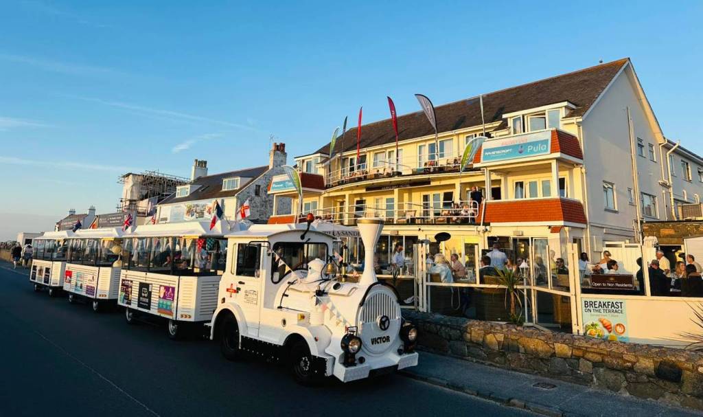 A white tourist road train is parked along a seafront road in the early evening light, beside a busy restaurant with people dining on an outdoor terrace. The long train is decorated with small flags, and the buildings behind it glow warmly in the sunset.