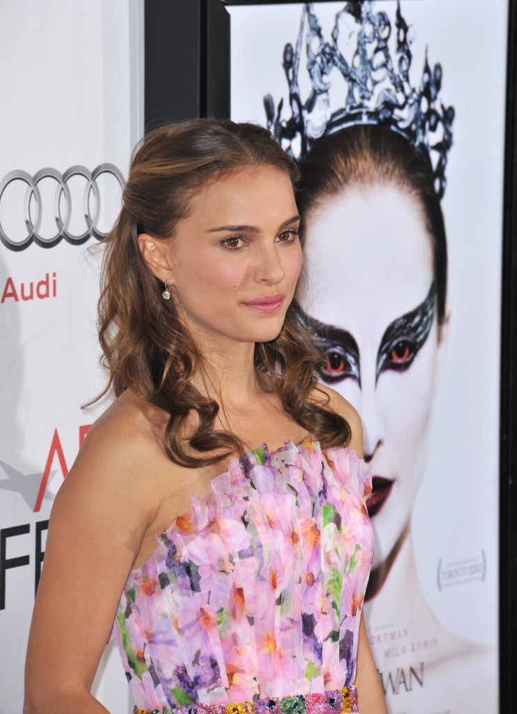 Natalie Portman. A woman with long dark hair wearing a strapless, multicolored floral-patterned gown poses in front of a large Black Swan movie poster at a red-carpet event. The poster behind the person features a dramatic close-up of a performer in stylized stage makeup with dark, wing‑shaped eye makeup and an ornate crown. Parts of an event backdrop with branding and logos are also visible around the poster.