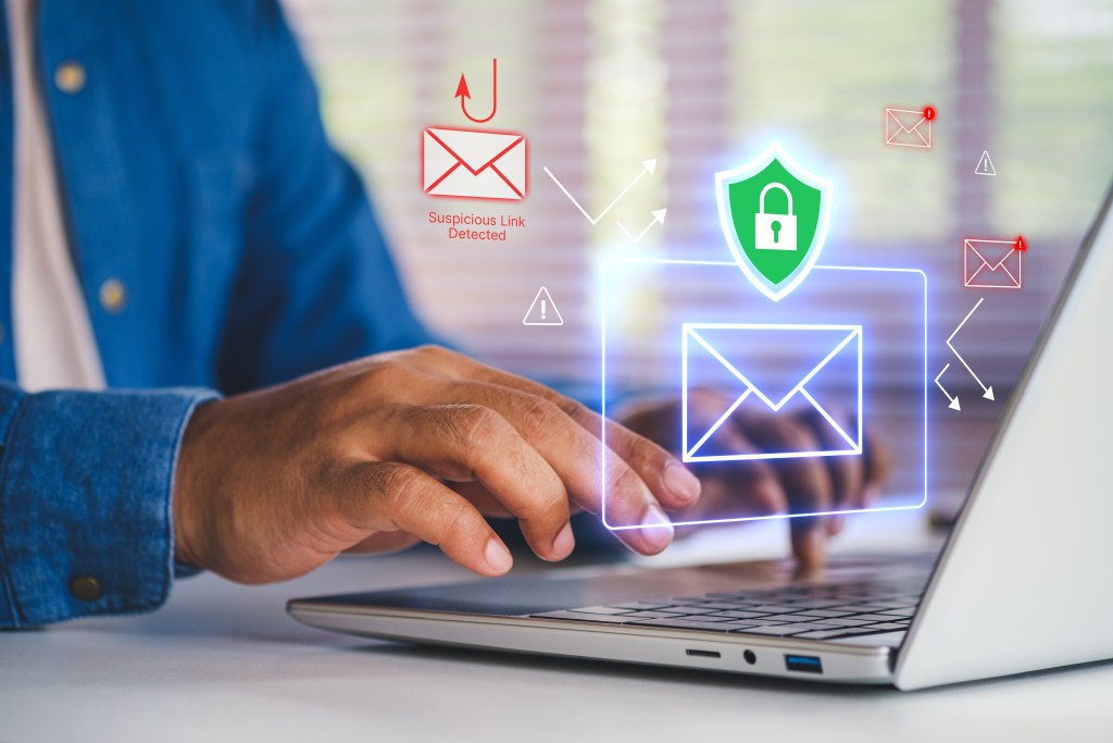 A person types on a laptop while digital graphics of email icons, a green security shield, and red warning symbols appear above the keyboard, representing phishing threats and secure email protection.