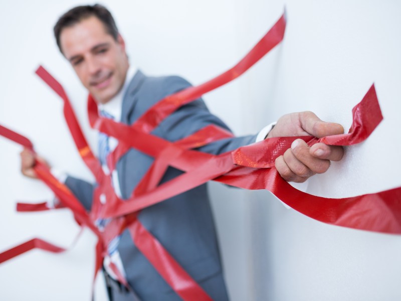 A businessman in a suit, stuck to a wall by red tape. He's struggling to get out of it.