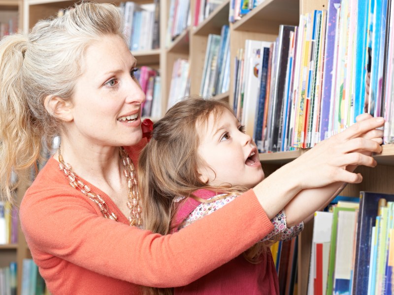 An mother and daughter stand close together in a library aisle, reaching toward a shelf filled with colorful books. The woman, wearing a coral-colored top and layered necklaces, extends an arm toward the books while the child, dressed in patterned clothing, reaches upward beside them. Rows of bookshelves filled with various titles line the background.