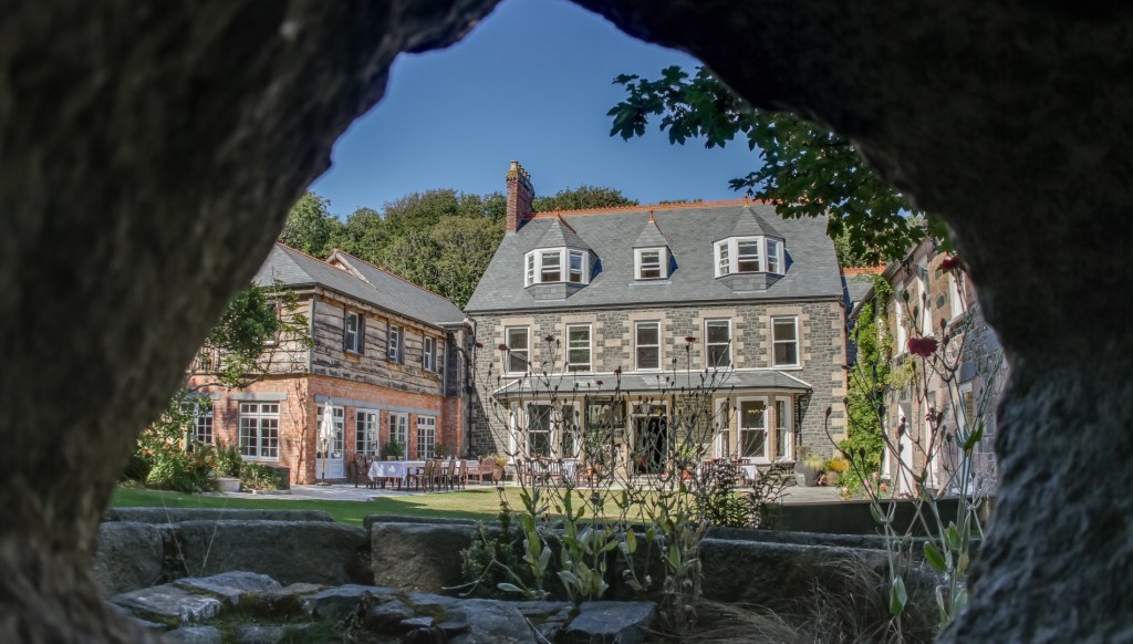 A large stone and brick hotel with dormer windows is viewed through a natural stone opening, with a lawn, plants and outdoor seating in the garden in front.
