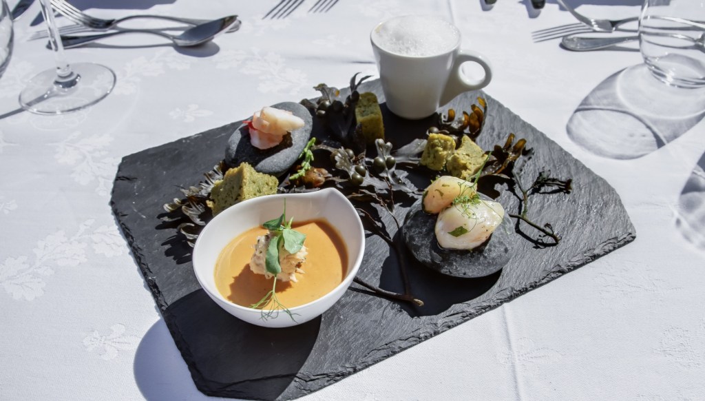 A selection of seafood and small savoury bites is arranged on a black slate plate, including prawns, seaweed, a small bowl of orange soup and a foamy drink in a white cup, set on a white tablecloth.
