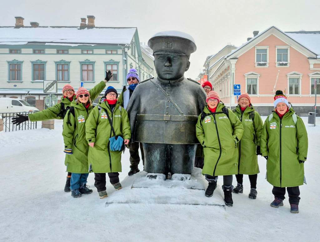 A group wearing matching green coats stands around a large metal statue in a snowy town square lined with colourful old buildings.