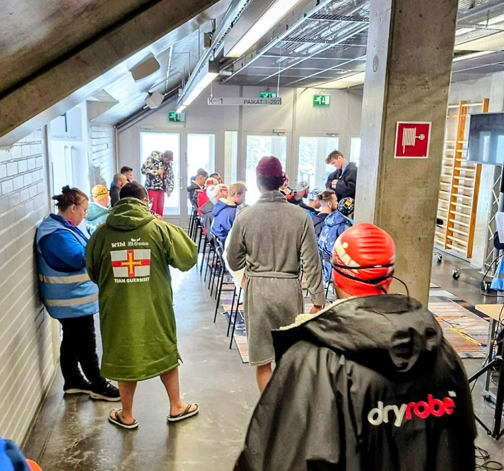 Competitors in robes and winter gear sit and gather inside a concrete building during an event briefing; one robe shows a Guernsey team logo.