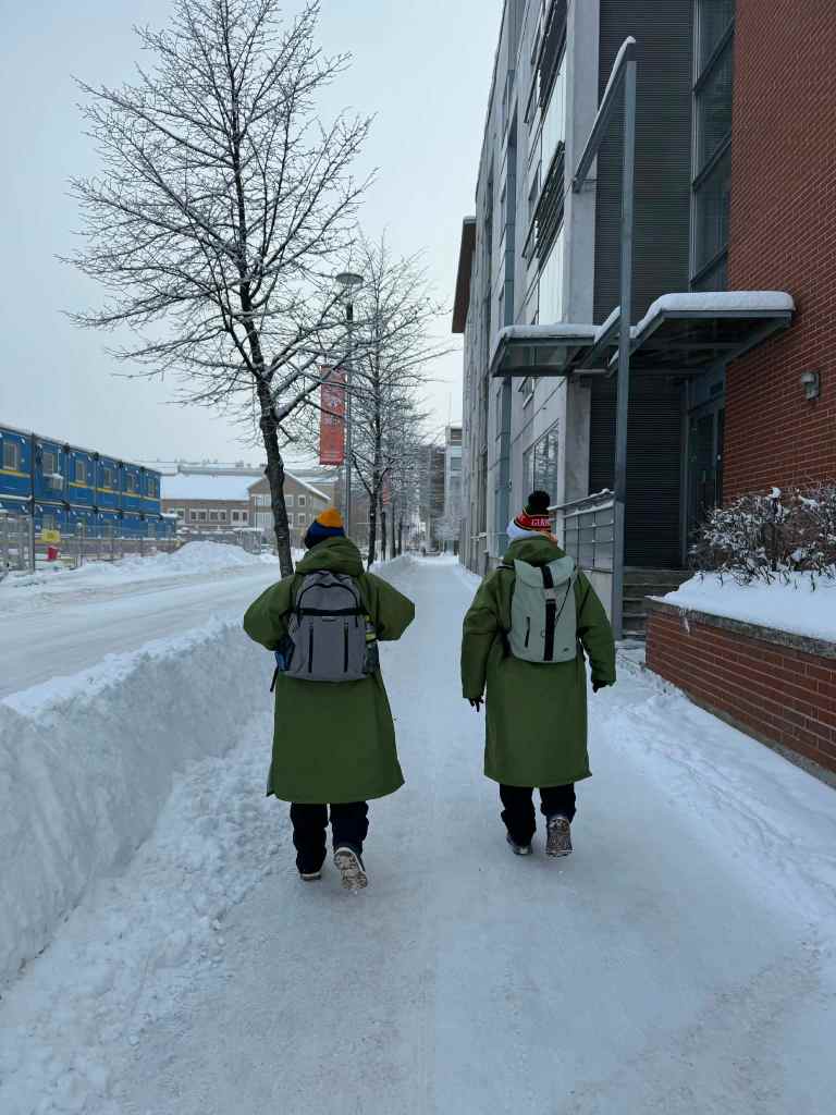 Two people in long green coats and backpacks walk along a snowy pavement beside buildings and bare trees.