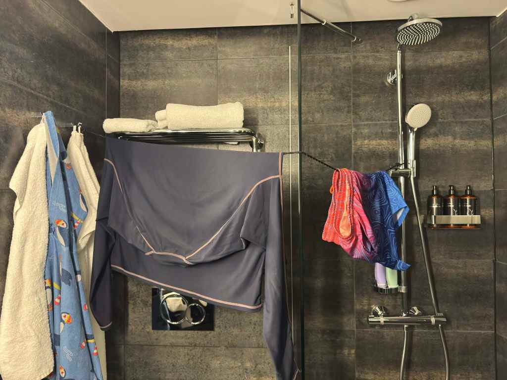 A bathroom with dark tiled walls, where towels and clothes hang to dry from hooks, a rack, and a shower rail above a modern shower unit.